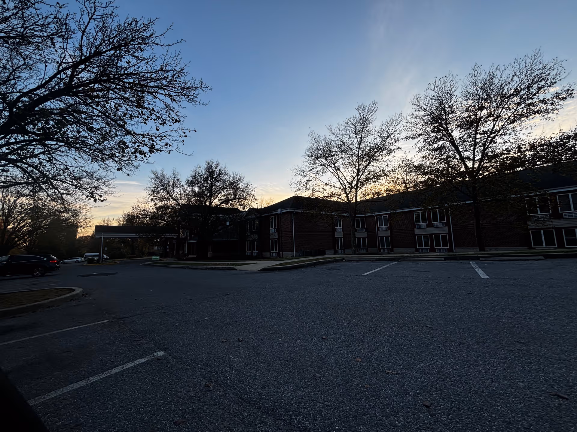 Parking lot in front of a two-story brick building with several leafless trees around, under a clear sky at sunset.