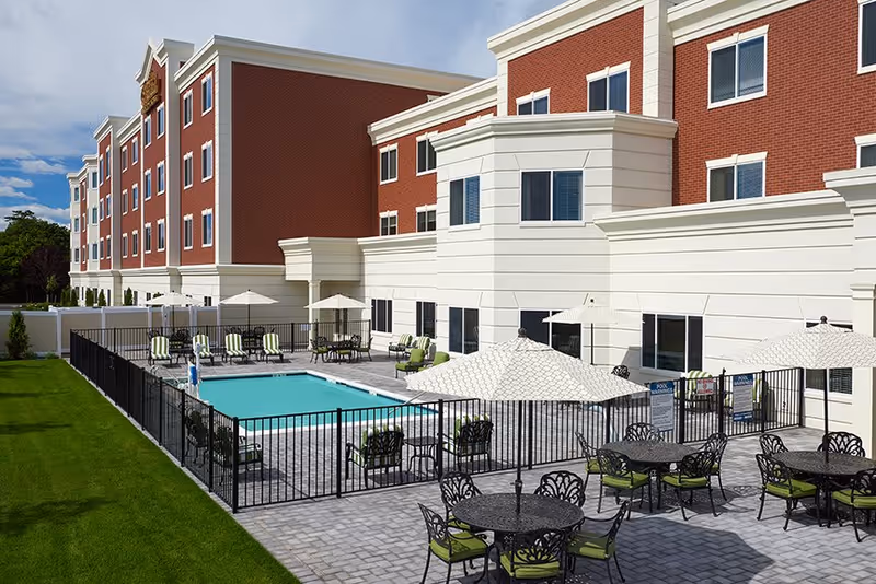 Outdoor patio area with a swimming pool surrounded by a black metal fence. Several round tables with green cushioned chairs and large white umbrellas are arranged on the paved patio. The background shows a multi-story brick and cream-colored building under a partly cloudy sky.