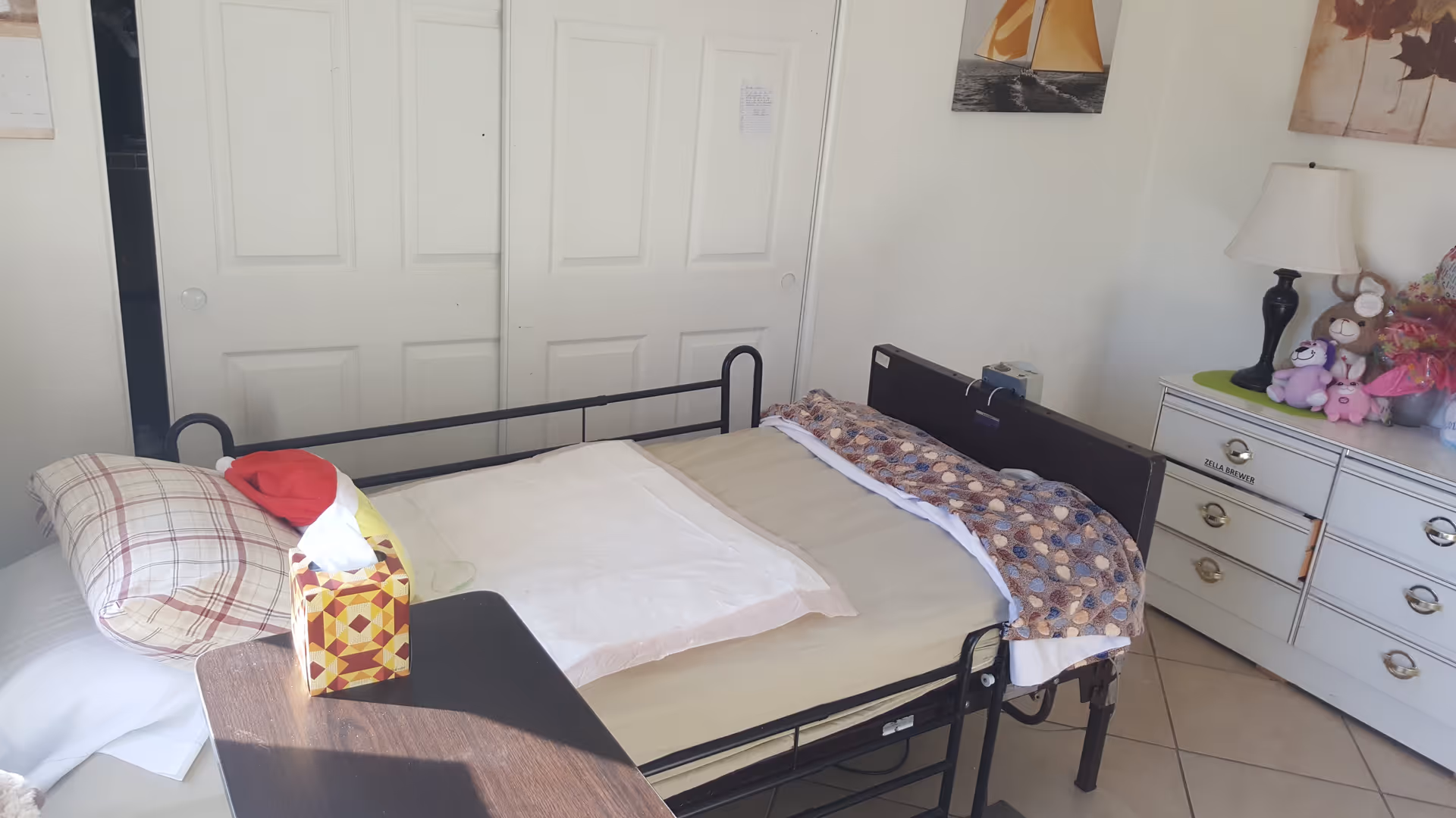 A single bed with a metal frame in a room, featuring a beige bedspread and a patterned blanket at the foot. There is a plaid pillow and a tissue box on a small wooden table next to the bed. A white dresser with drawers is visible on the right side, topped with a lamp, stuffed animals, and a bouquet of flowers. The background shows white sliding closet doors and a wall with a sailboat picture.