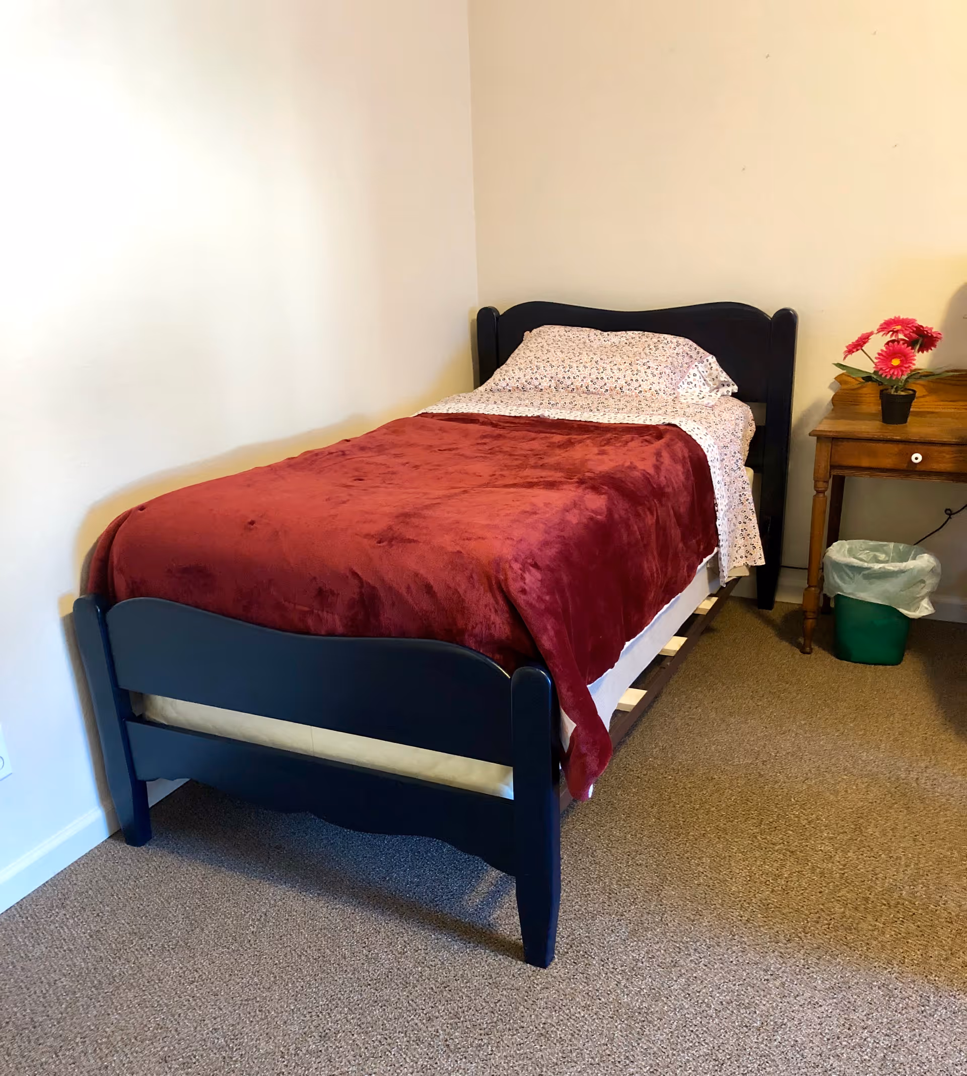 A single bed with a dark frame and burgundy blanket beside a wooden nightstand with a small vase of flowers in a simple bedroom.
