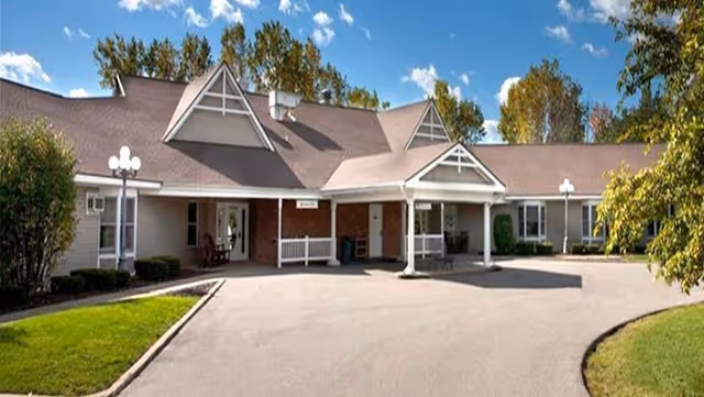 Front exterior view of Whispering Woods Retirement Village building with a covered entrance, surrounded by green lawns and trees under a blue sky with some clouds.
