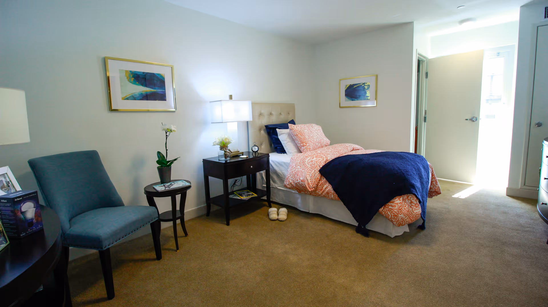 A neatly arranged bedroom with a single bed featuring orange and white patterned bedding and a dark blue throw blanket. There is a nightstand with a lamp, clock, and flower vase next to the bed. A blue upholstered chair and a small round table with a potted plant are positioned against the wall. Two framed abstract artworks hang on the walls. The room has beige carpeting and an open door letting in natural light.