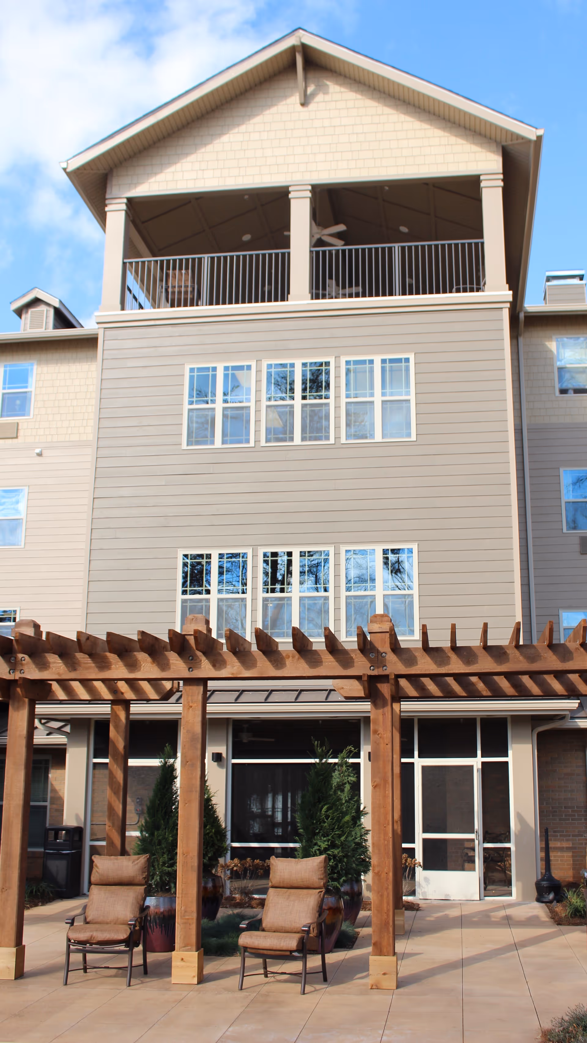 Exterior view of a multi-story senior living facility with beige siding and multiple windows. The building features a covered balcony on the top floor with ceiling fans. In the foreground, there is a wooden pergola structure with two cushioned chairs underneath, surrounded by potted plants on a paved patio area.