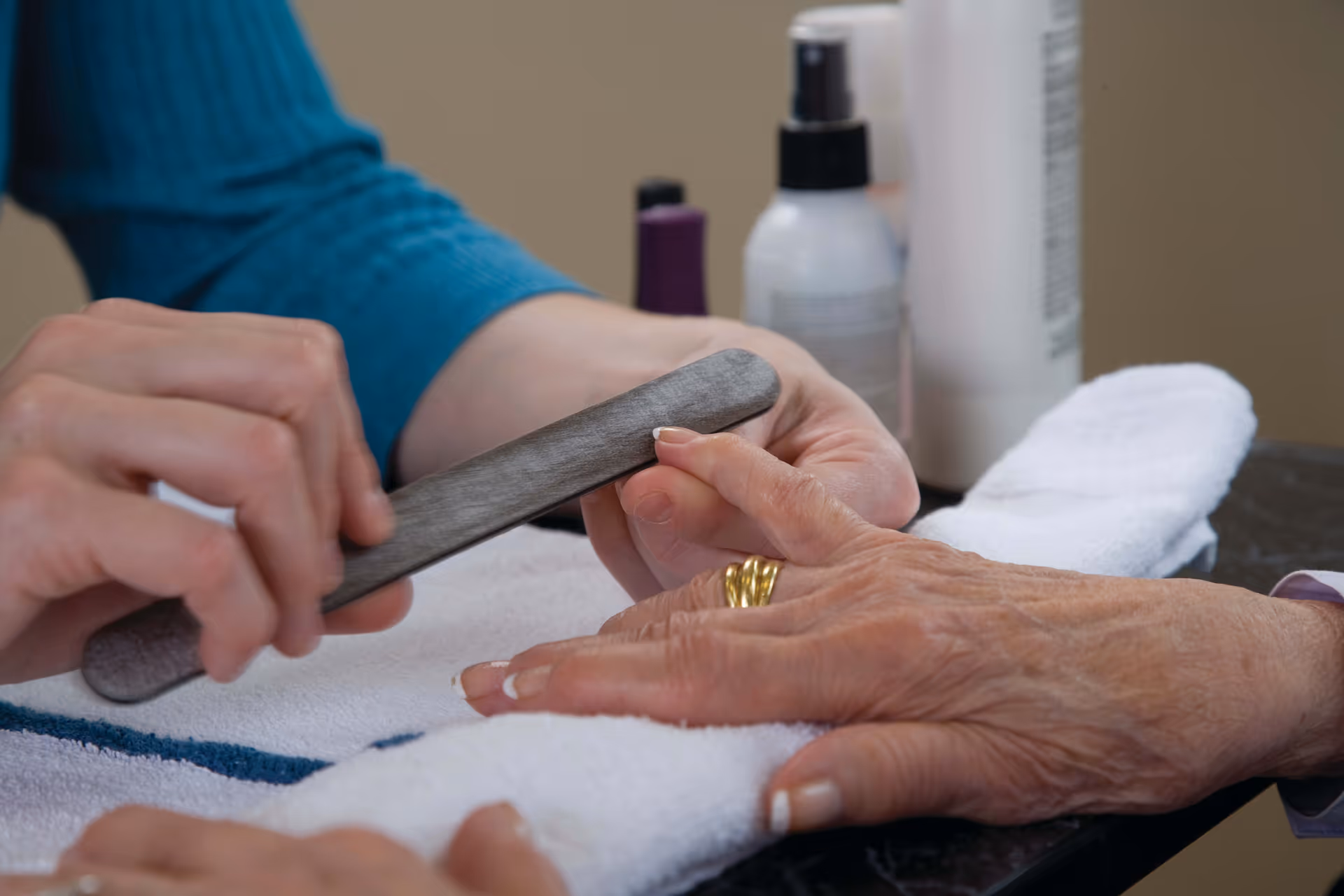 A close-up of a person filing the fingernails of an elderly person wearing a gold ring, with nail care products in the background on a table covered with a white towel.