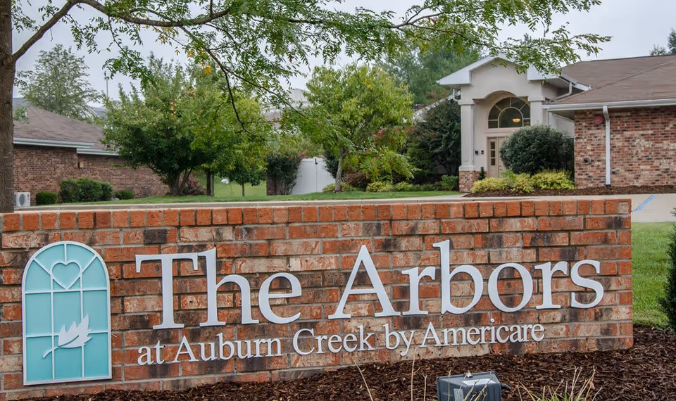 A brick sign outside a senior living facility that reads 'The Arbors at Auburn Creek by Americare' with a decorative window and leaf logo on the left. Behind the sign, there are trees, shrubs, and a building entrance with a large window and door.