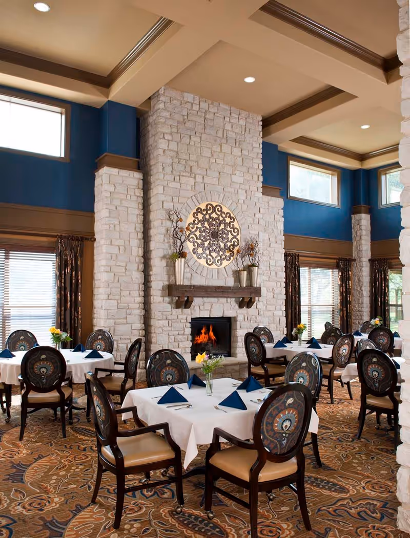 Dining room with multiple tables covered in white tablecloths, each set with blue folded napkins and small flower vases. The room features a large stone fireplace with a decorative metal wall art above it, blue walls with large windows, and patterned carpet flooring.