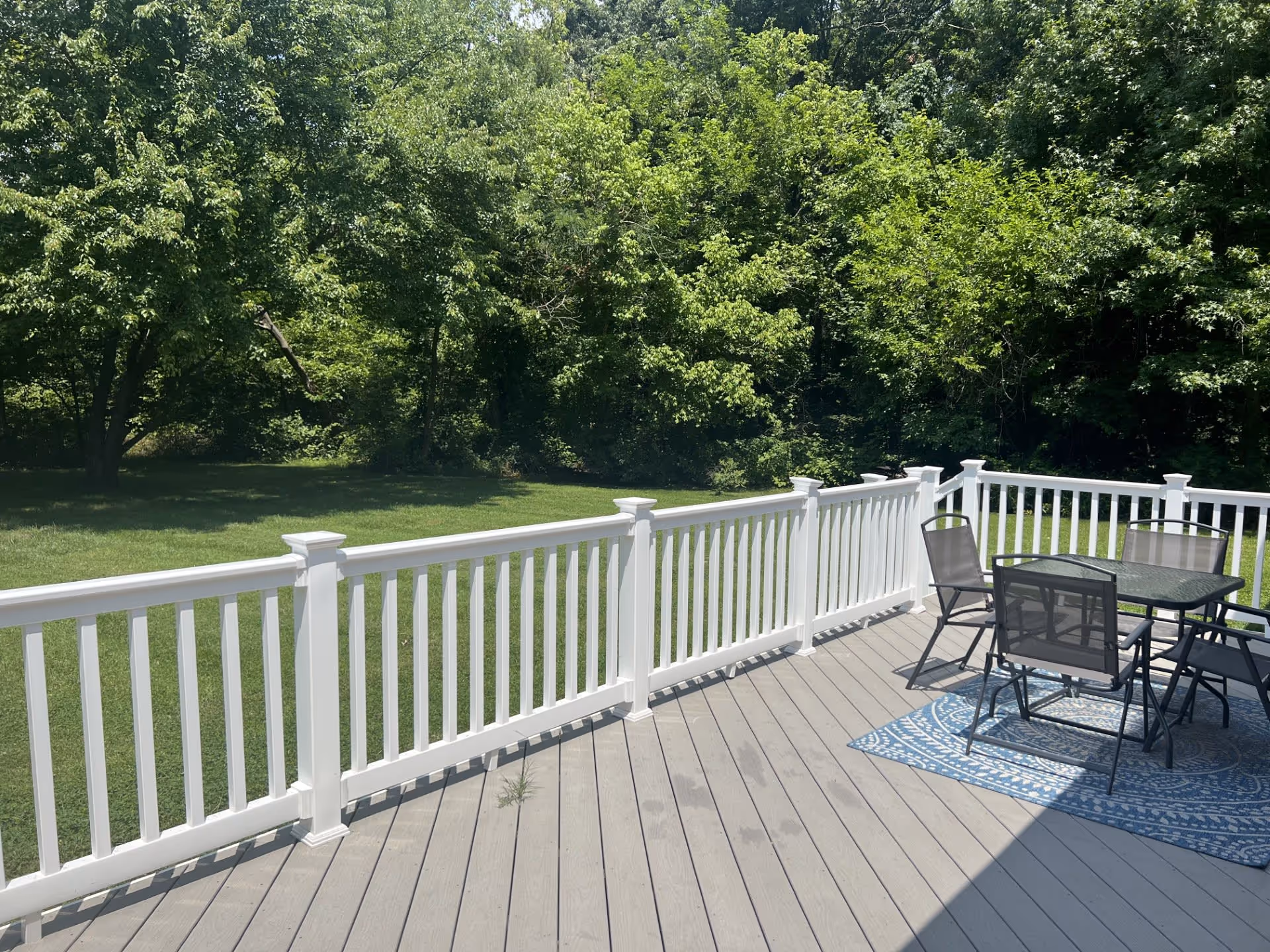 Outdoor deck area with gray wooden flooring, white railing, and a glass-top table with four chairs on a blue patterned rug. The deck overlooks a green grassy yard with dense trees and foliage in the background under bright sunlight.