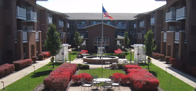 View of a courtyard in a senior living facility with a central fountain, surrounded by red bushes and green grass. There are tables and chairs arranged near the fountain, and an American flag on a flagpole in the center. The courtyard is flanked by multi-story residential buildings with balconies.