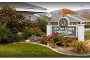 Entrance sign reading 'Life Care Center' amid landscaped flowers and shrubs in front of a single-story care building with mountains and autumn trees behind.
