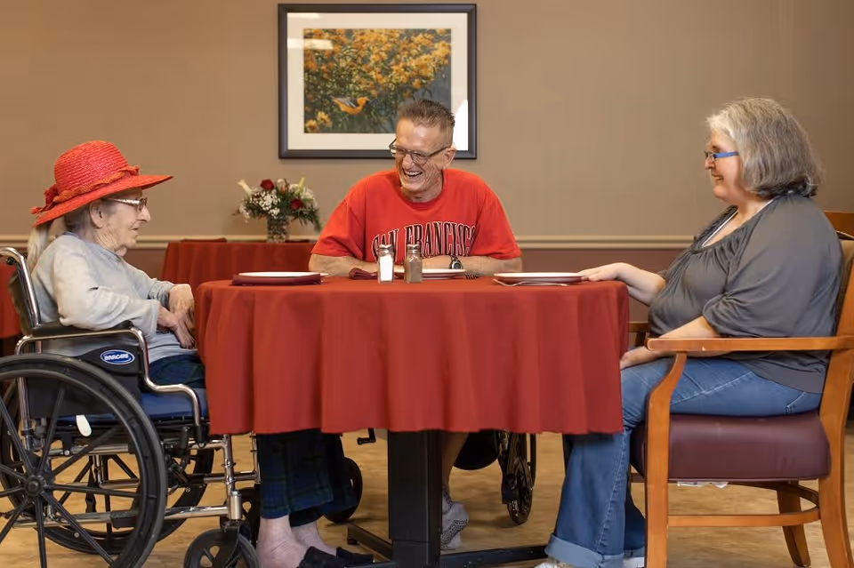 Three elderly individuals sitting around a round table with a red tablecloth in a dining area. One woman in a wheelchair wearing a red hat and glasses, a man in a red San Francisco t-shirt smiling, and another woman with gray hair and glasses sitting on a chair. A framed picture of a bird and flowers hangs on the wall behind them.