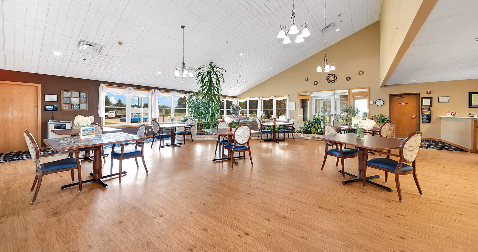 A spacious, well-lit dining area with several wooden tables and chairs arranged neatly. The room features large windows with white curtains, allowing natural light to fill the space. There are plants placed around the room, including a tall plant in the center. The walls are painted in warm tones, and there are decorative elements on the walls. The ceiling has multiple hanging light fixtures, and the floor is covered with wood-like flooring.