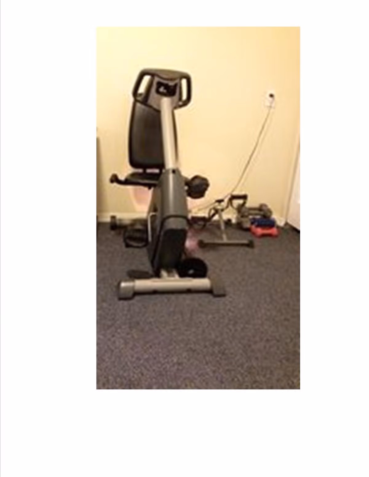 A small indoor exercise area with a recumbent stationary bike and some free weights on a carpeted floor against a beige wall.