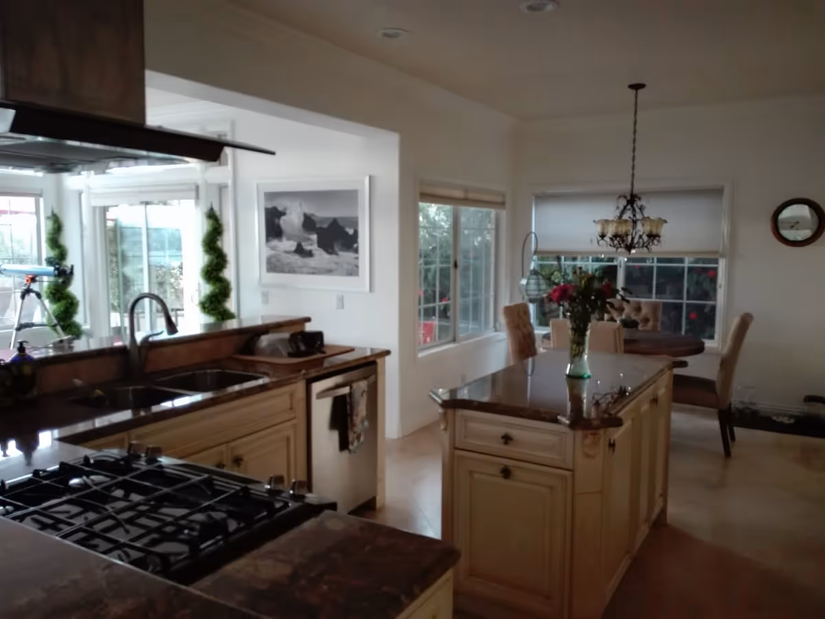 Interior view of a kitchen and dining area with a gas stove, sink, and kitchen island with a vase of flowers. The dining area has a round table with chairs and a chandelier hanging above it. Large windows provide natural light and a view of greenery outside.