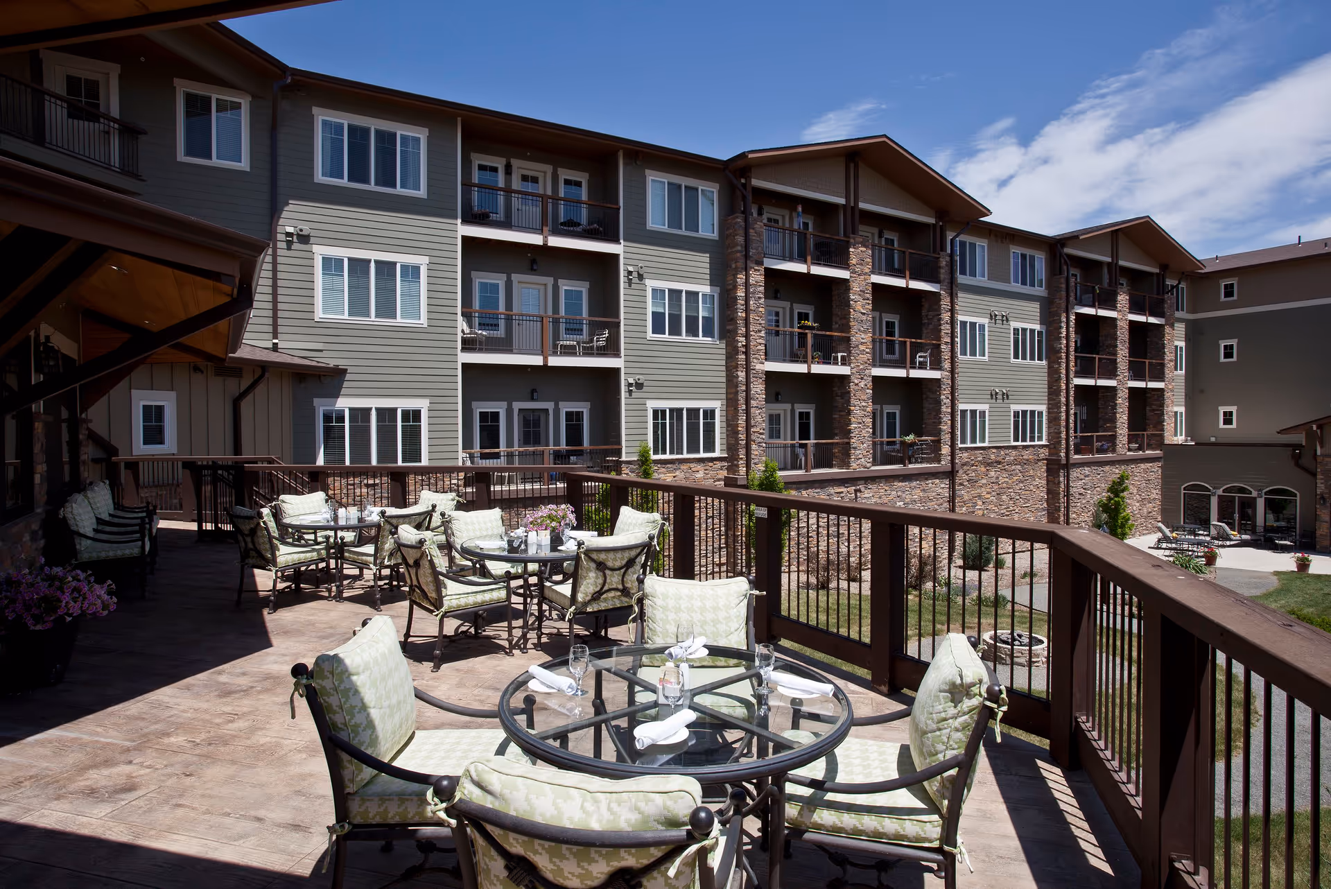 Outdoor patio area with several glass-top tables and cushioned chairs arranged for dining or socializing. The patio overlooks a multi-story residential building with balconies and large windows under a partly cloudy blue sky.
