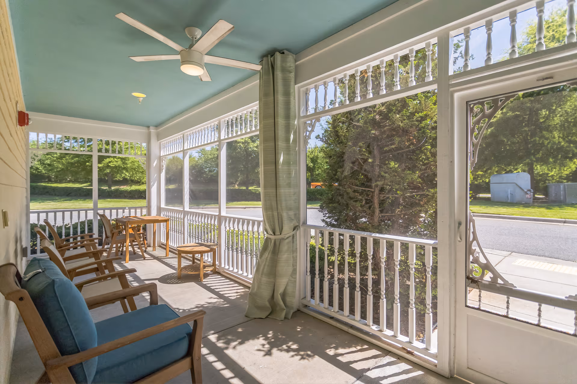 A screened-in porch area with several wooden chairs with blue cushions, a small wooden table, and a ceiling fan. The porch overlooks a green outdoor area with trees and a road visible through the screen panels.