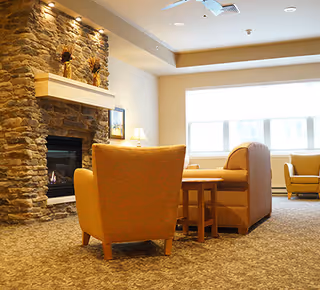 Cozy living room area with a stone fireplace, a lit fire, and comfortable upholstered chairs and sofas arranged around a wooden coffee table. A window with blinds allows natural light into the room, and a table lamp is placed on a side table near the fireplace.