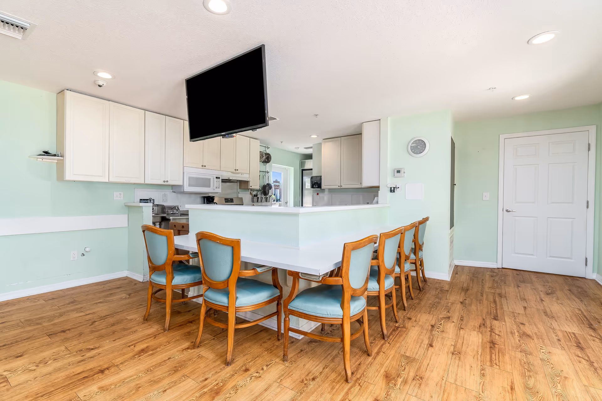 A bright kitchen and dining area with light green walls and wooden flooring. The kitchen features white cabinets, a microwave, and a stove. A white countertop extends into a dining bar with six wooden chairs upholstered in light blue fabric. A flat-screen TV is mounted above the dining bar. A white door and a wall clock are visible in the background.