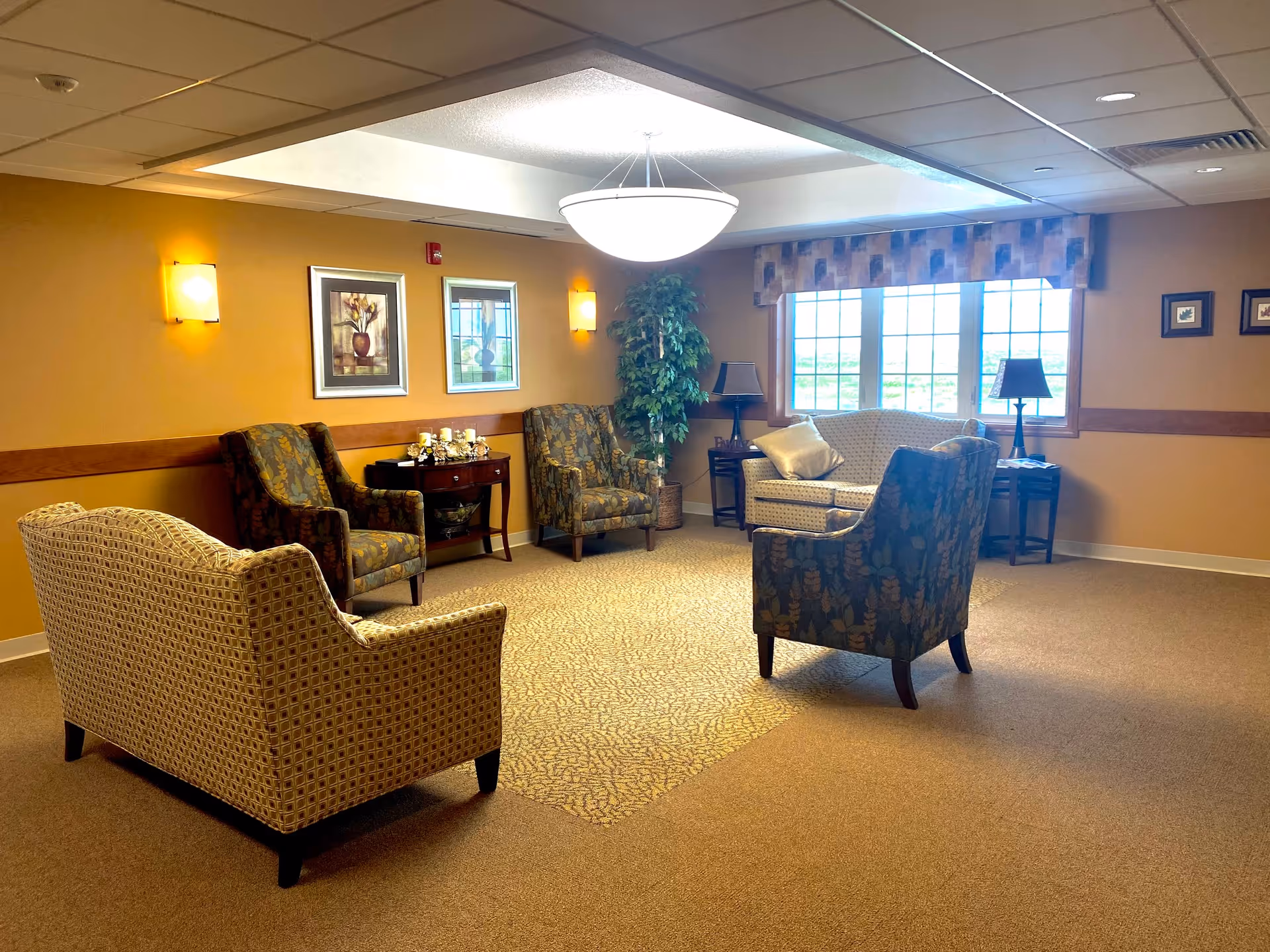 A cozy sitting area in a senior living facility with several upholstered armchairs and a loveseat arranged around a small table. The room has warm yellow walls, framed artwork, a large window with patterned valance, two table lamps, and a ceiling light fixture. There is a potted plant in the corner and carpeted flooring.