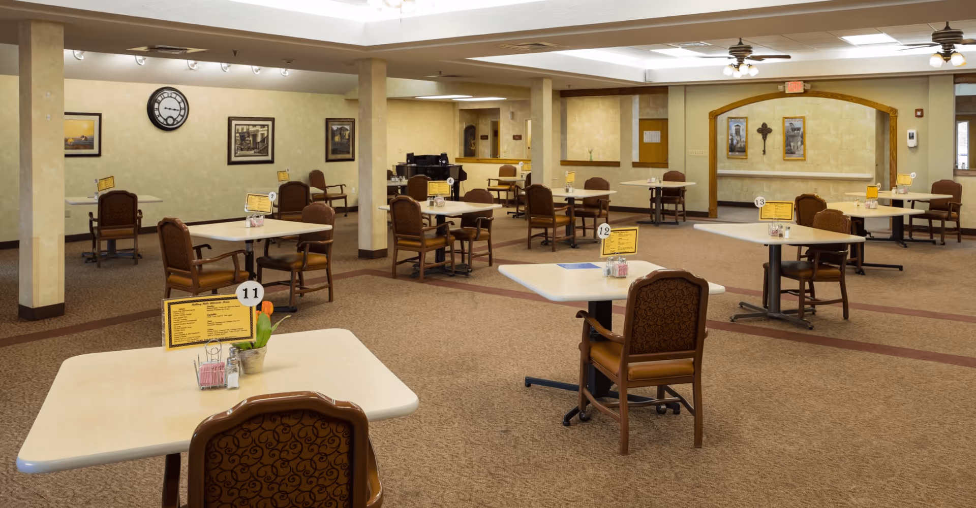 Spacious, well-lit assisted living dining room with multiple small tables and chairs arranged across a carpeted floor.