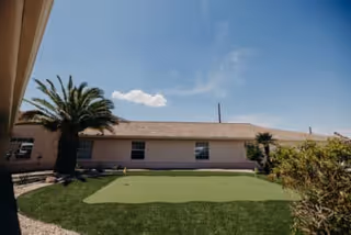 Outdoor area of a senior living facility with a putting green, palm trees, shrubs, and a single-story building under a clear blue sky.