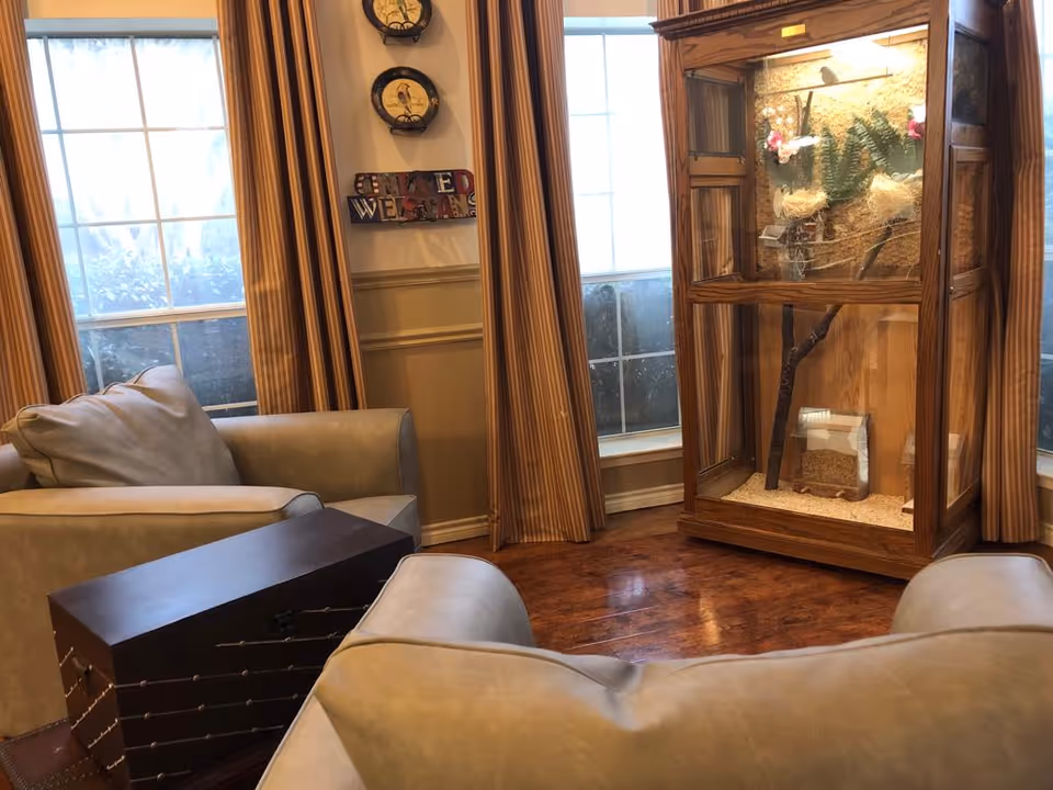 A cozy sitting area with two beige armchairs and a dark wooden chest-style coffee table between them. Behind the chairs are two large windows with striped curtains. On the right side, there is a wooden display case with a bird habitat inside. The walls are decorated with two round wall clocks and a sign that reads 'GOD BLESS OUR VETERANS'.