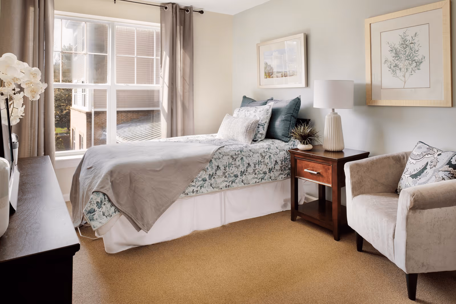 A cozy bedroom with a neatly made bed featuring floral bedding and multiple pillows. Next to the bed is a wooden nightstand with a white table lamp and a small plant. A comfortable beige armchair with a patterned cushion is positioned nearby. The room has beige carpeting, light-colored walls with framed artwork, and a large window with beige curtains letting in natural light.