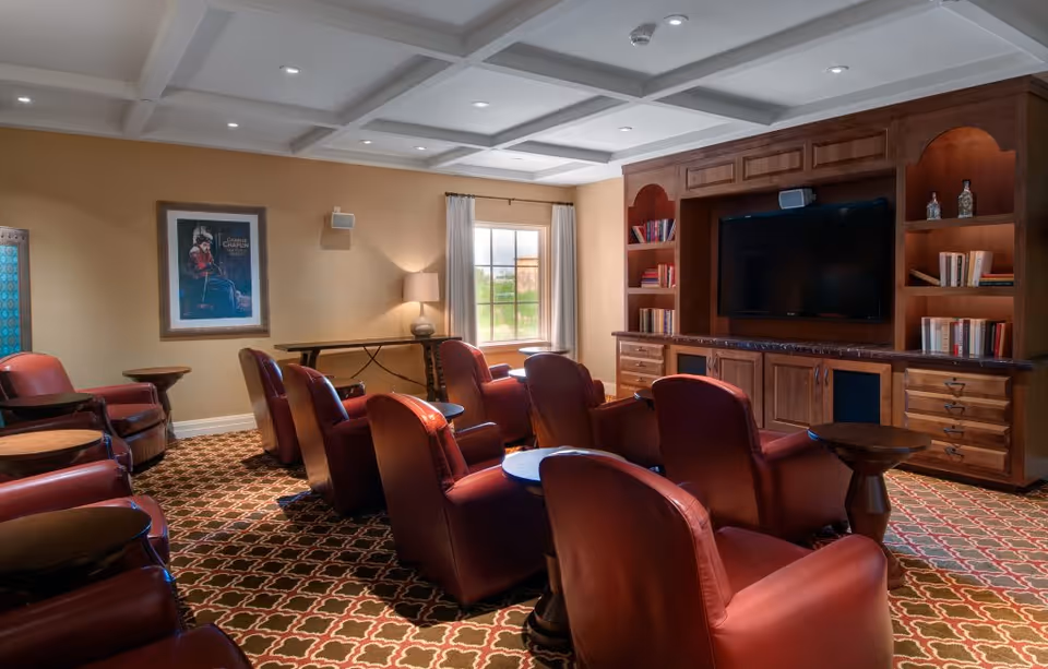 A cozy media room with several red leather recliners arranged in rows facing a large flat-screen TV mounted on a wooden built-in cabinet with shelves filled with books and decorative items. The room has a patterned carpet, beige walls, a window with curtains letting in natural light, and a framed Charlie Chaplin poster on one wall.