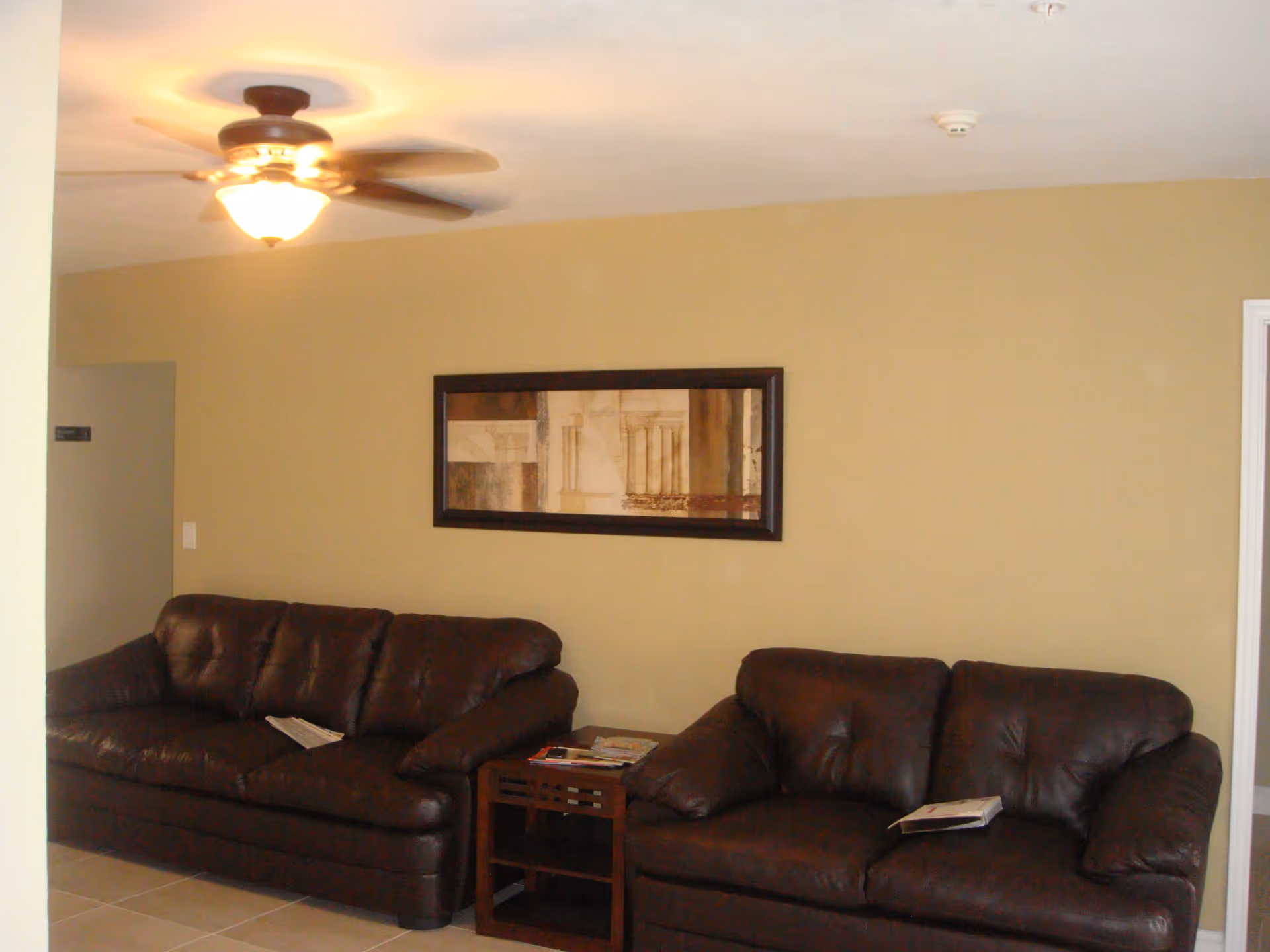Interior view of a living room area with two dark brown leather sofas separated by a small wooden side table holding magazines. A framed abstract artwork hangs on the beige wall above the sofas. A ceiling fan with a light is turned on above.