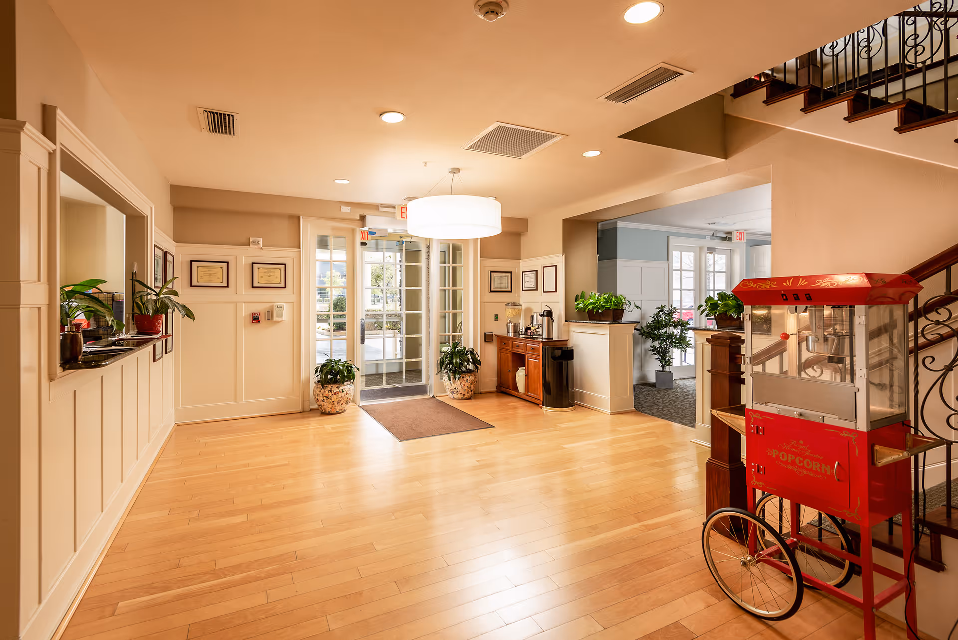 Bright and spacious lobby area with light wood flooring, a red vintage popcorn machine on wheels near a staircase, potted plants, a wooden cabinet with coffee and water dispensers, and glass double doors leading outside.