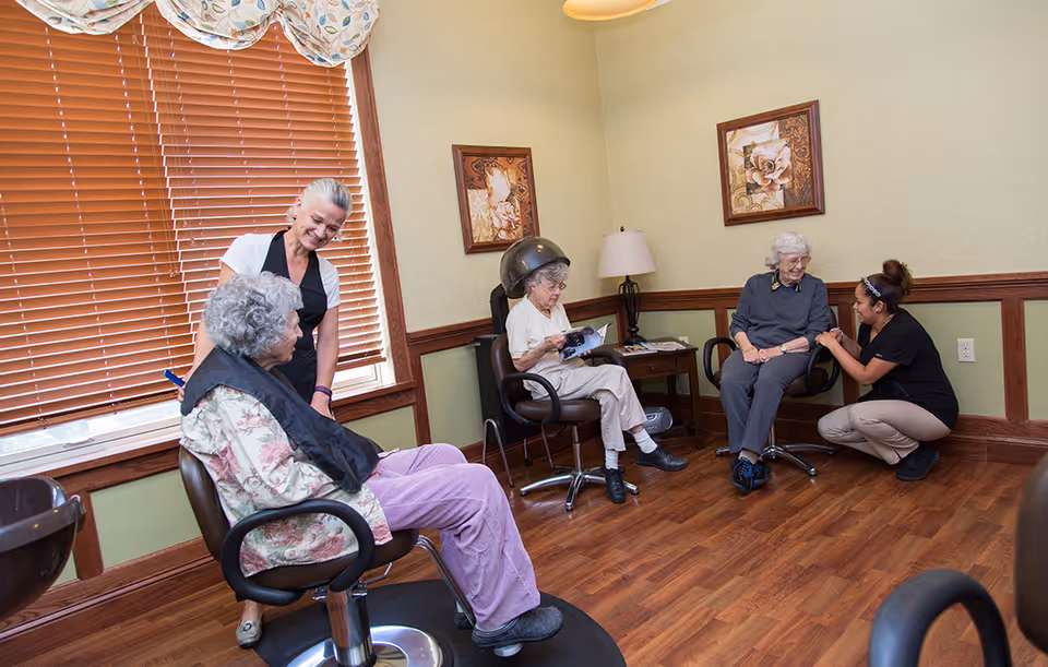 A room with three elderly women seated in salon chairs, one under a hair dryer reading a magazine, another having her hair attended to by a staff member, and a third seated near a window with blinds. The room has wooden flooring, light green walls with wood trim, two framed floral paintings, and a table with a lamp.