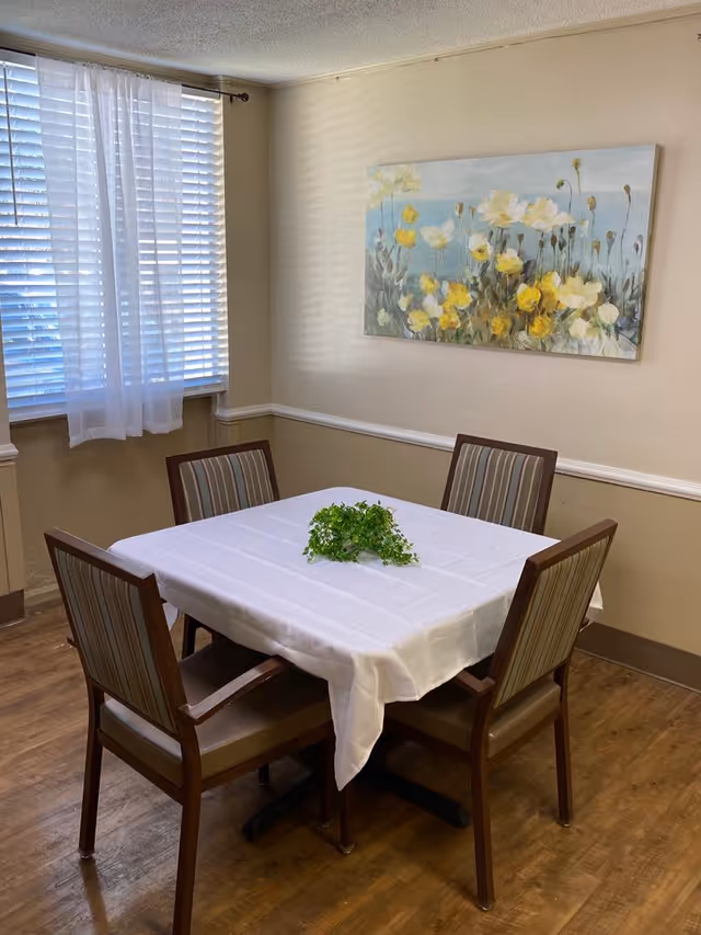 A small dining area with a square table covered with a white tablecloth and a small green plant centerpiece. Four wooden chairs with striped cushions surround the table. A window with white blinds and sheer curtains is on the left wall, and a painting of yellow and white flowers hangs on the beige wall behind the table.