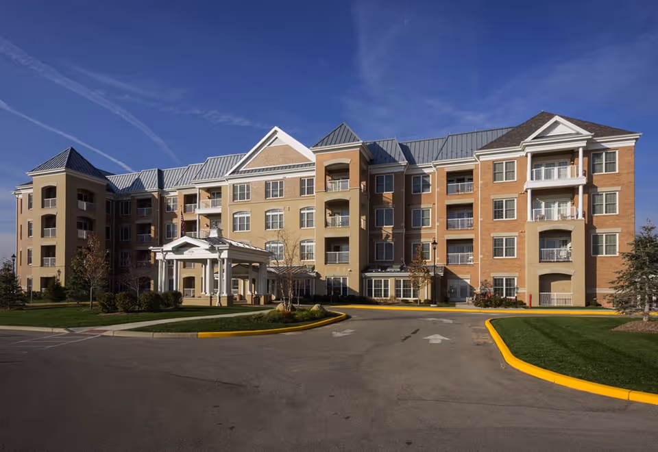 Exterior view of a multi-story senior living facility building with a covered entrance, balconies, and a driveway surrounded by landscaped grass and trees under a clear blue sky.
