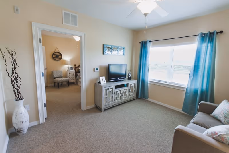 A bright living room with beige walls and carpet, featuring a gray sofa with patterned pillows on the right, a window with blue curtains letting in natural light, and a decorative TV stand with a flat-screen television. A doorway leads to a bedroom with a chair, nightstand, and bed visible.
