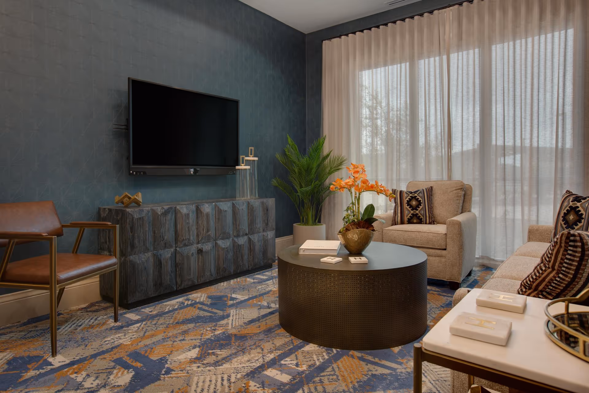 A cozy living room with a mounted flat-screen TV on a dark blue textured wall, a wooden cabinet below the TV, a brown leather chair, a round black coffee table with a flower vase and books, a beige armchair and sofa with patterned cushions, a side table with decorative items, and large windows covered with sheer curtains letting in natural light.