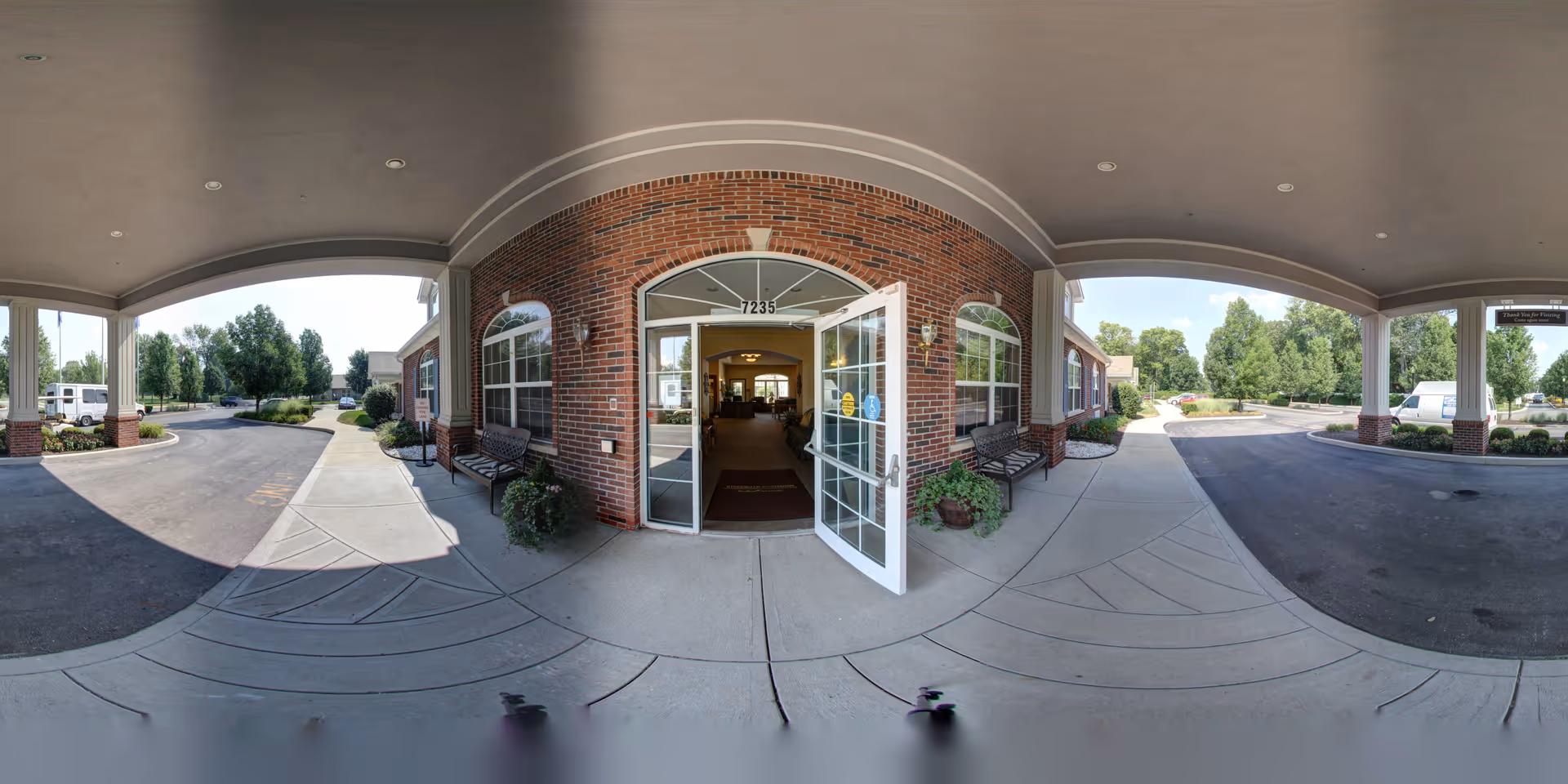 Covered entrance area of Five Star Residences of Noblesville showing a brick building with an open glass door leading inside. There are benches and potted plants on either side of the entrance. The driveway and sidewalk curve around the entrance with trees and greenery in the background under a clear sky.