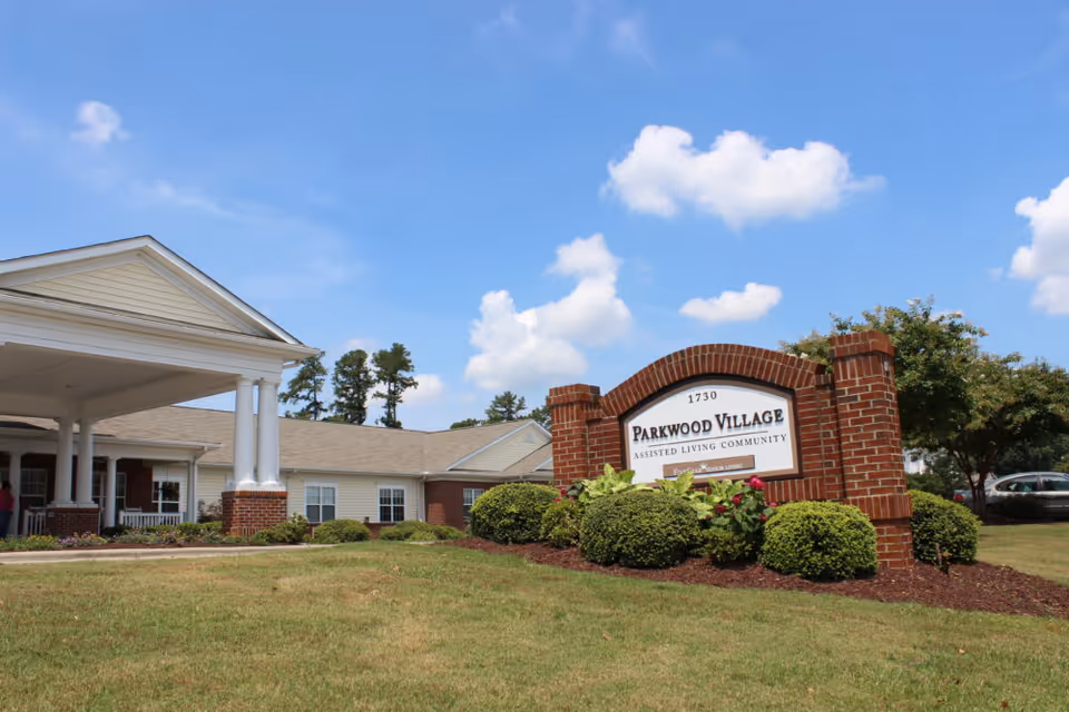 Brick entrance sign reading 'Parkwood Village' in front of a one-story assisted living building with a covered portico under a blue sky.