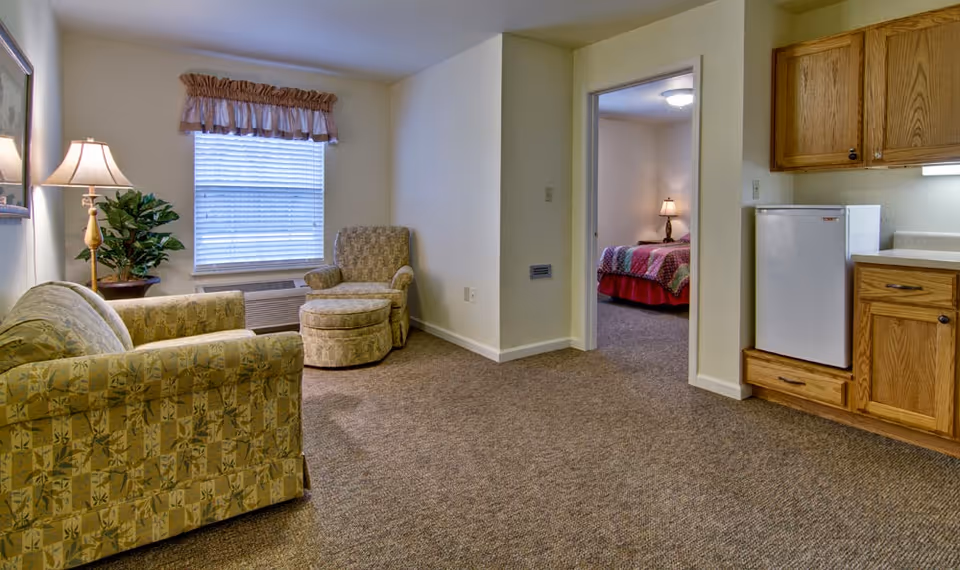 A cozy living area in a senior living facility with a patterned armchair and matching ottoman, a sofa, a floor lamp, and a potted plant near a window with blinds and a valance. To the right, there is a small kitchenette with wooden cabinets and a mini refrigerator. A doorway leads to a bedroom with a bed covered in a colorful quilt and a bedside lamp.