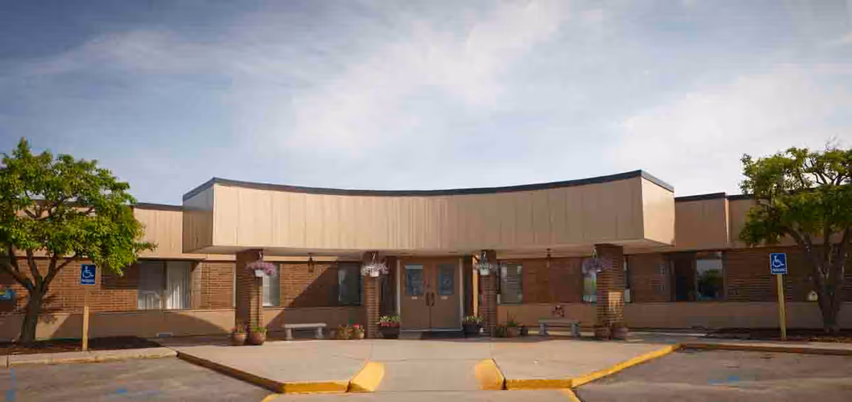Front entrance of a single-story brick senior living facility with a curved canopy, parking area, and handicap signs.