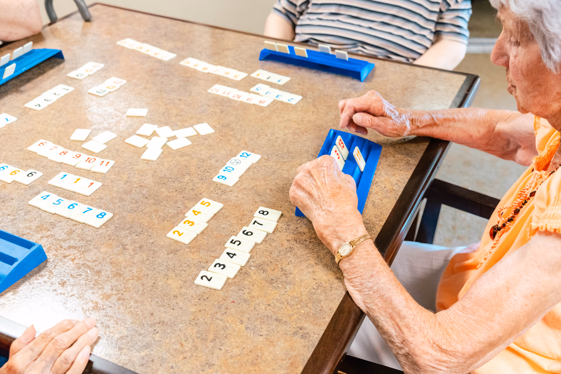 Elderly people sitting around a table playing a tile-based game, with one woman in an orange shirt holding tiles on a blue rack.