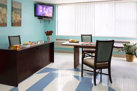 Bright interior common room with tables and chairs, a wall-mounted TV, and games on the table.
