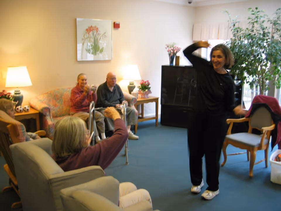 A group of elderly people sitting in a living room area with a woman standing and engaging with them. The room has comfortable chairs, a floral couch, lamps, a large TV, and plants, creating a warm and inviting atmosphere.