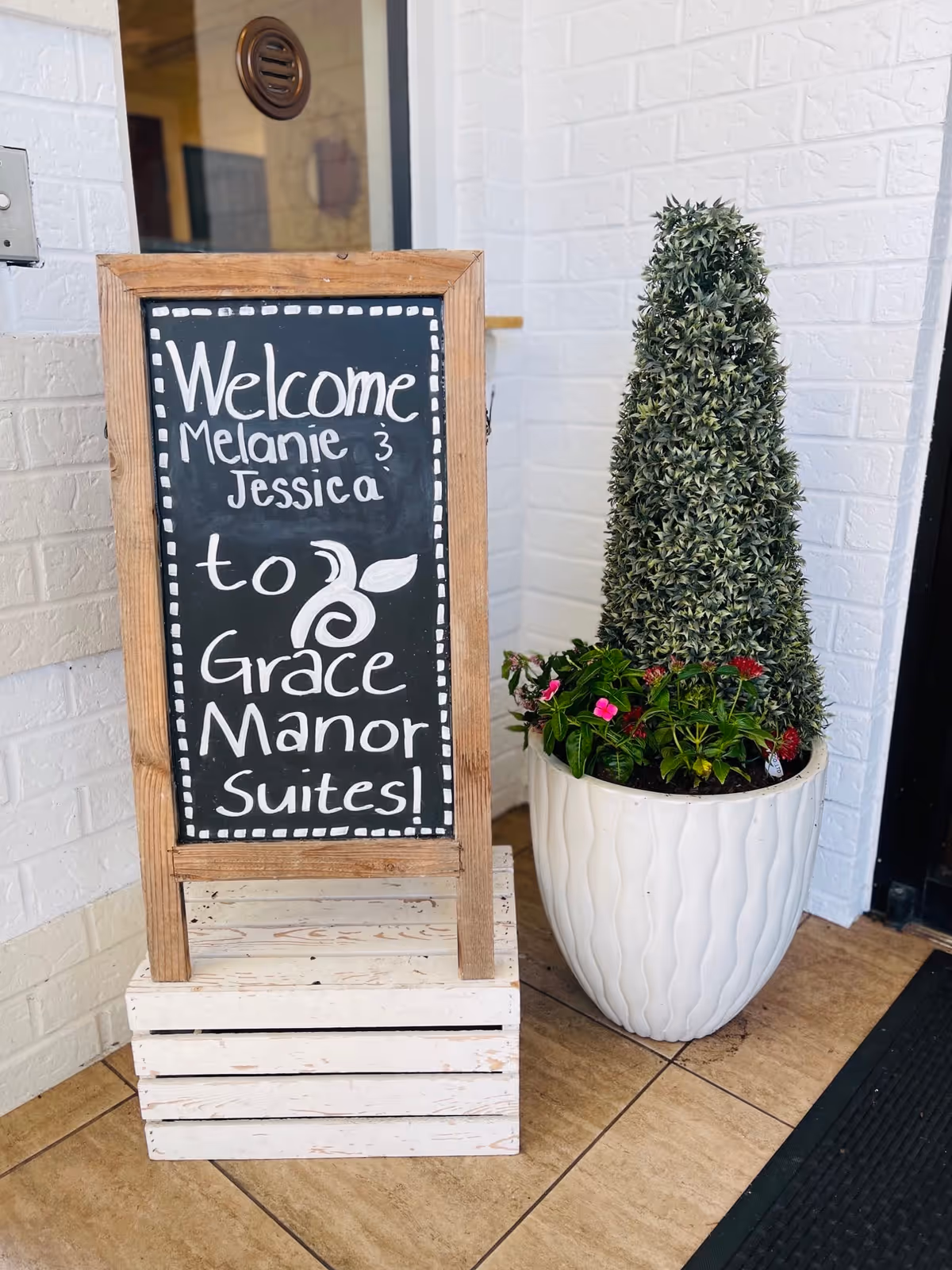 A wooden-framed chalkboard sign on a white crate welcomes Melanie and Jessica to Grace Manor Suites, placed next to a tall potted plant with green foliage and small flowers, set against a white brick wall near a glass door.