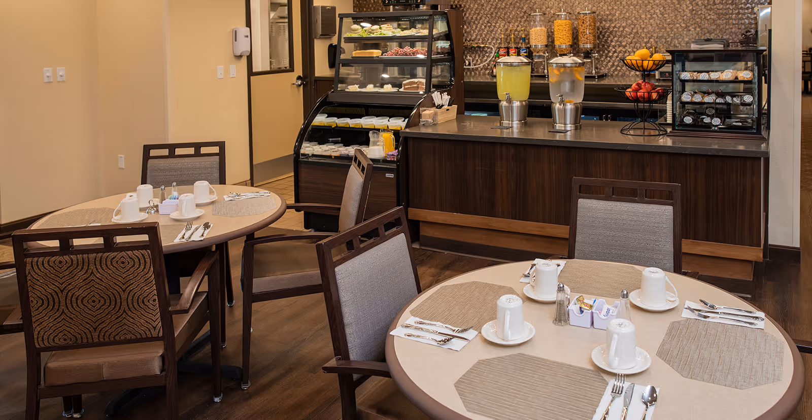 Dining area with round tables set with cups, plates, and cutlery. In the background, there is a counter with beverage dispensers containing lemonade and water, a fruit stand with apples and oranges, and display cases with various food items. The room has wooden flooring and neutral-colored walls.