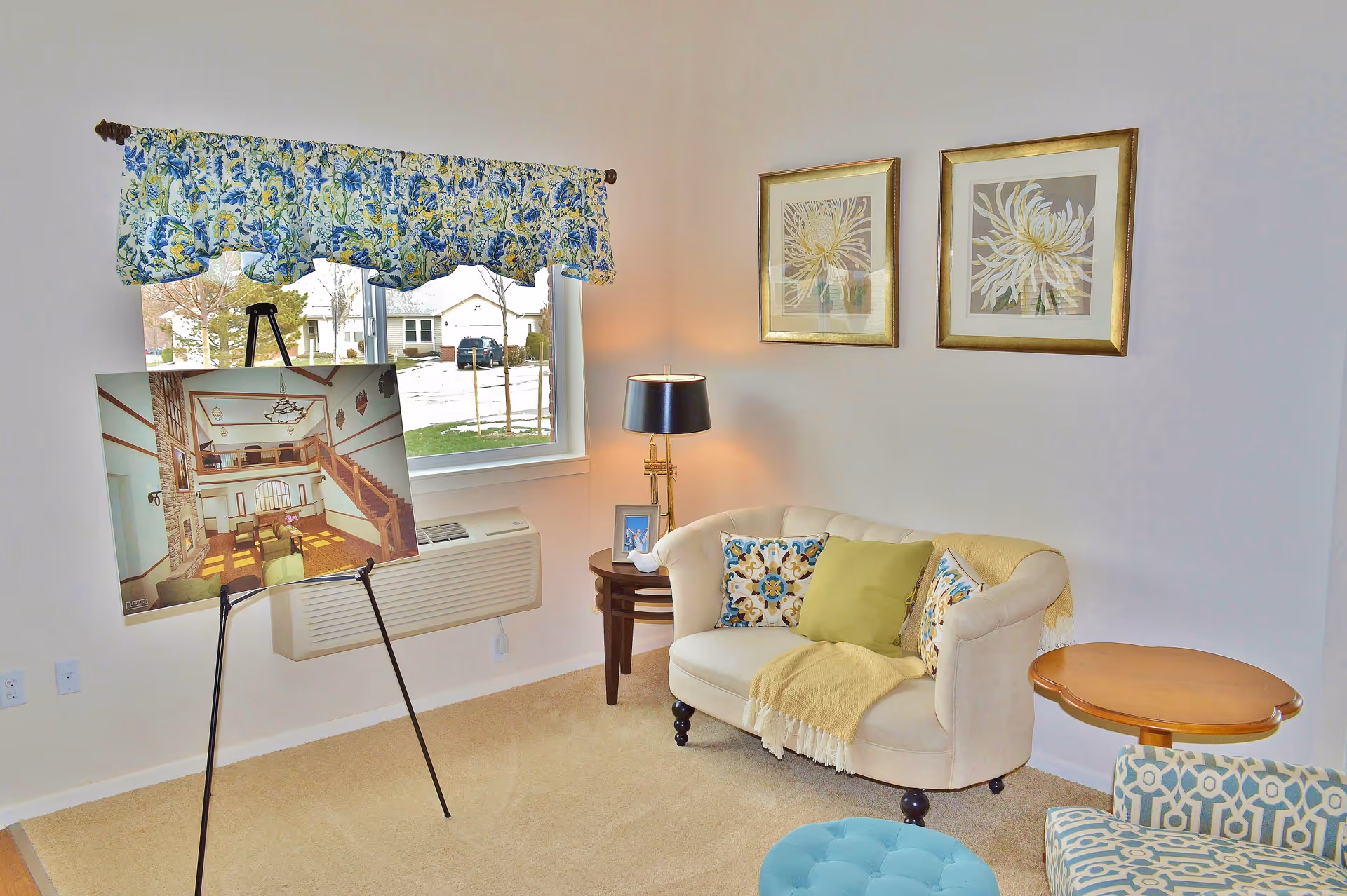 A cozy living room with a small beige loveseat adorned with colorful pillows and a yellow throw blanket. There is a round wooden side table next to the loveseat and a patterned armchair partially visible. A black lamp with a gold base sits on a small round table beside the loveseat. Two framed floral artworks hang on the wall above the loveseat. A window with a floral valance lets in natural light, and an easel holds a large photo of an interior space. The room has beige carpet and light-colored walls.