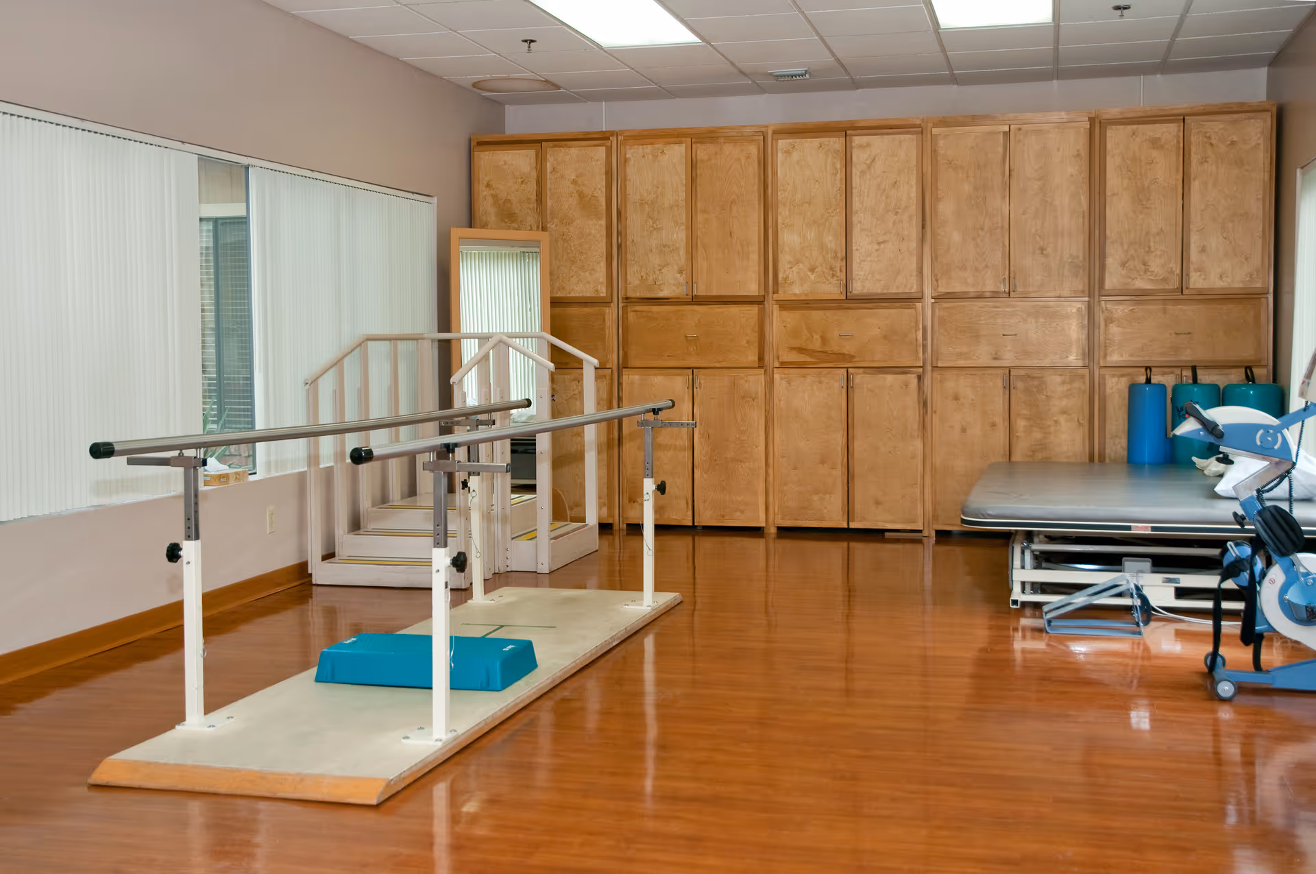 Physical therapy room with parallel bars for walking exercises, a set of stairs with handrails, a padded therapy table, wooden cabinets along the back wall, and a large mirror on one side.