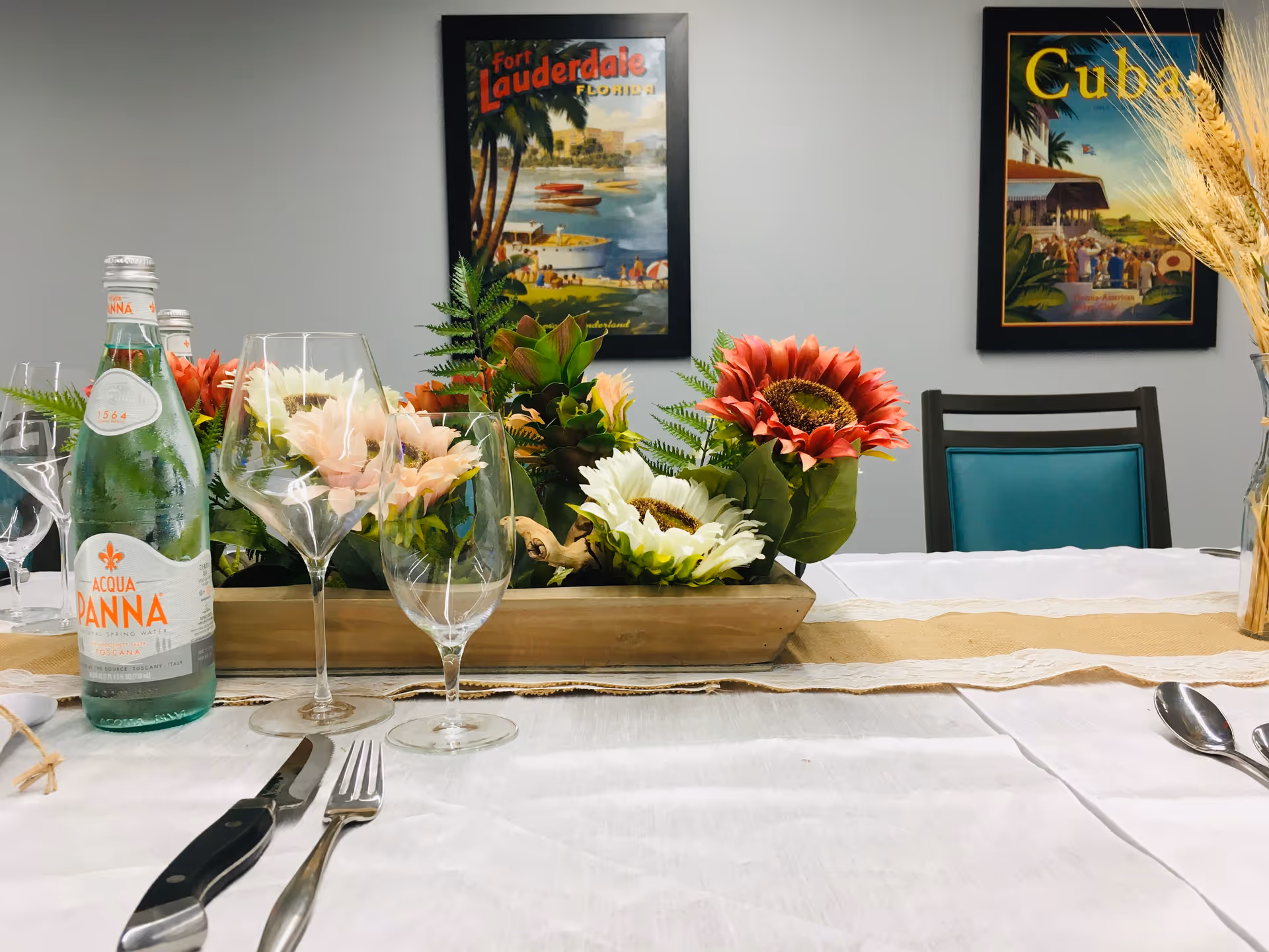 A dining table set with a white tablecloth, silverware, wine glasses, a bottle of Acqua Panna water, and a centerpiece of artificial flowers. Two framed vintage travel posters, one of Fort Lauderdale, Florida, and the other of Cuba, hang on the gray wall behind the table. A chair with a teal cushion is partially visible.