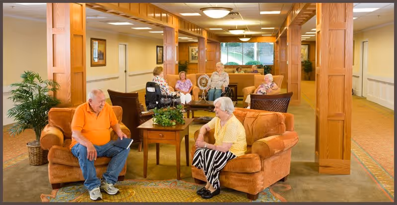 A spacious senior living facility common area with several elderly residents seated and engaged in conversation. The room features warm lighting, wooden columns, comfortable armchairs, and a central table with a plant. Large windows in the background allow natural light to fill the space.