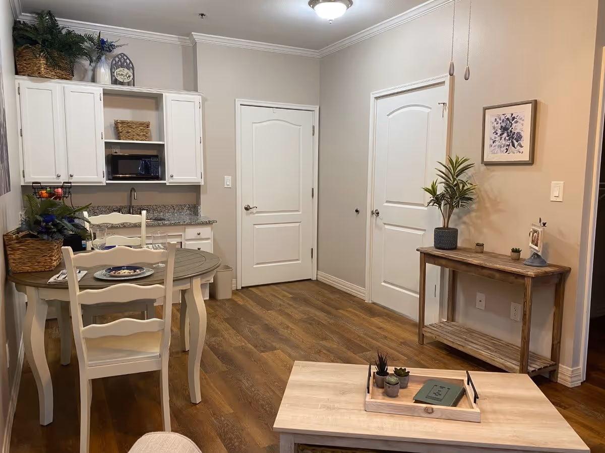 A cozy interior space featuring a small dining area with a round wooden table set for two, white chairs, and a kitchenette with white cabinets, a microwave, and a granite countertop. The room has wood flooring, two white doors, a wooden console table with a potted plant and small decor items, and a framed floral artwork on the wall.