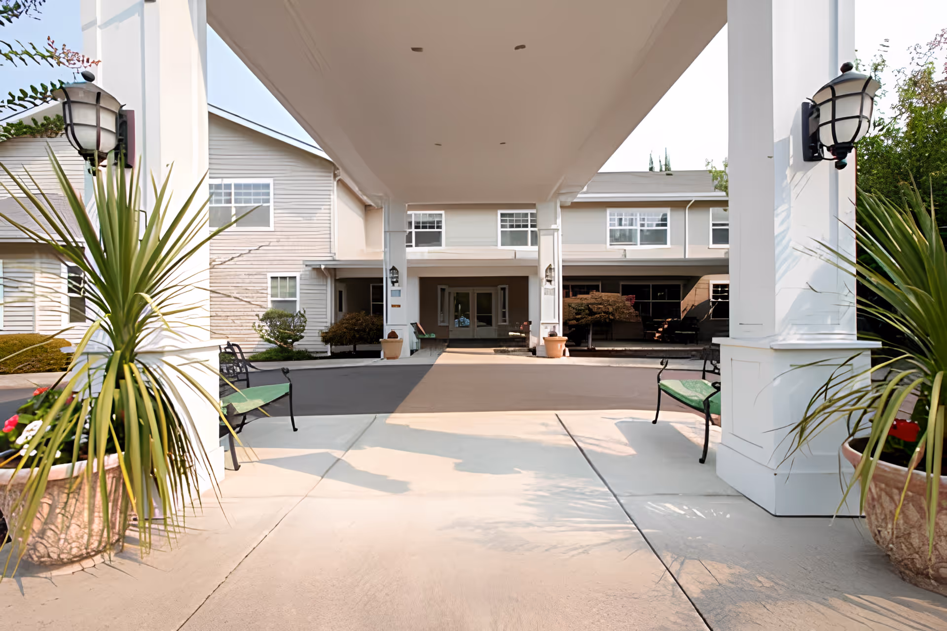 Entrance area of Oak Park Senior Living by Cogir showing a covered driveway with white pillars, benches on either side, large potted plants, and the front facade of the building in the background.