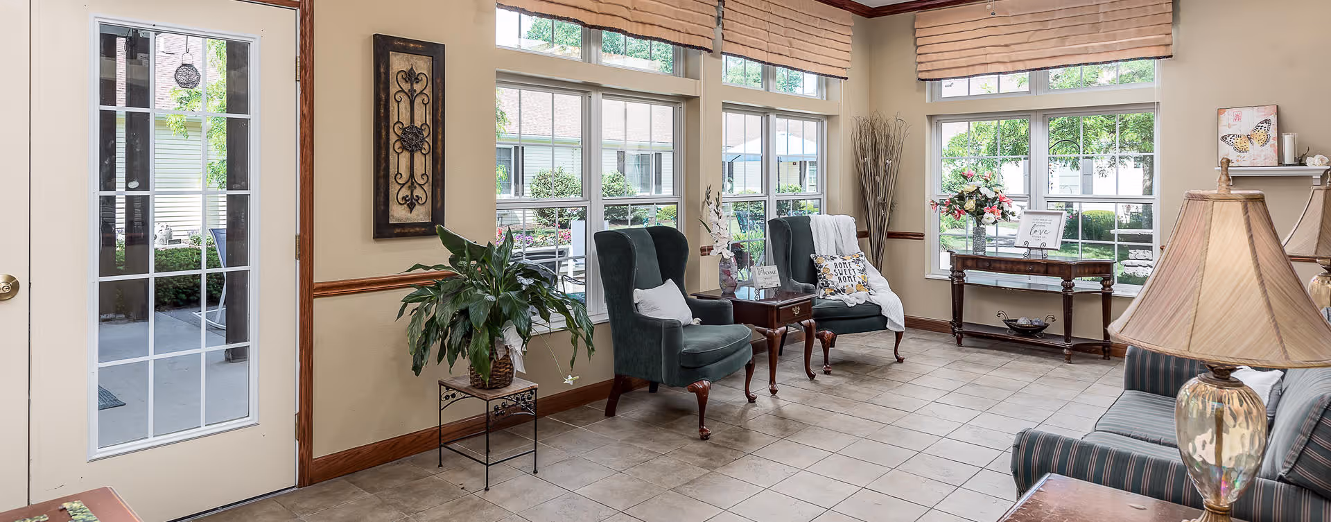 A bright and inviting living room area with large windows letting in natural light. The room features two green upholstered armchairs with pillows and a throw blanket, a small wooden side table between them, a striped sofa, a table lamp with a beige shade, a potted plant on a metal stand, and a wooden console table with decorative items including flowers and framed art. The floor is tiled, and there is a glass door leading outside.